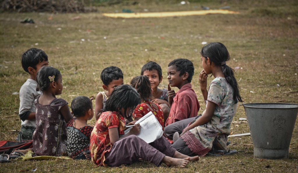faceless-girl-heartily-doing-exercise-in-book-with-pen-near-playing-indian-children-1440388 Group of full body ethnic children sitting on lawn and spending time together while unrecognizable little girl diligently doing homework wring in sheet of paper on street