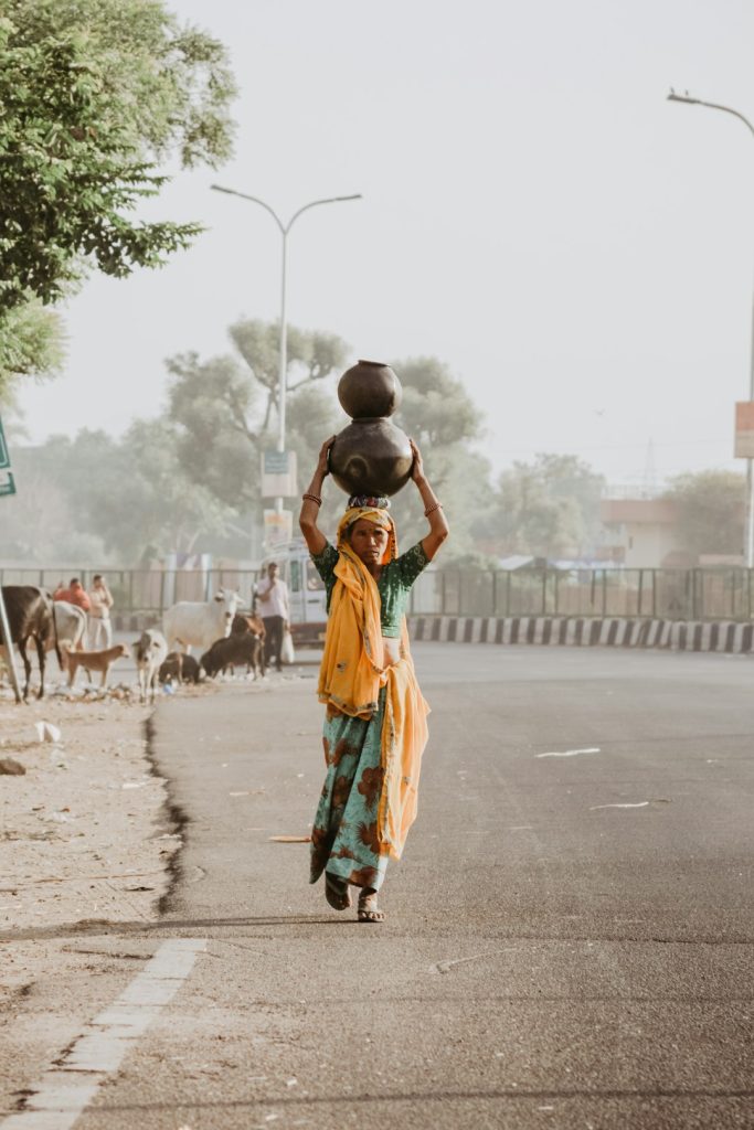 woman-walking-on-road-carrying-two-jars-on-head-olvv-o45gia A woman carries water home on her head